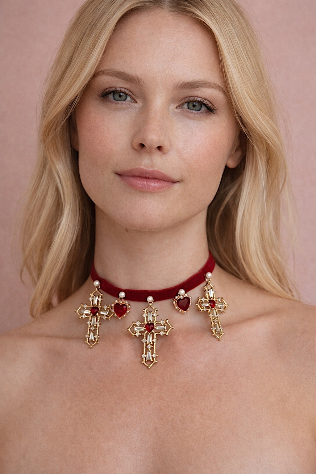 Blonde model wearing the Cross My Heart Jeweled Charm Bridal Choker with red velvet ribbon, gold cross charms, jeweled hearts, and pearls against a blush backdrop