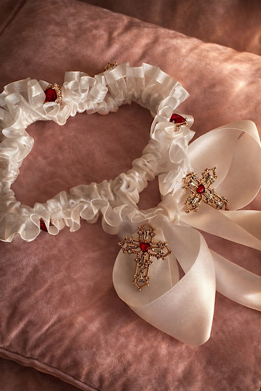 Cross My Heart Jeweled Charm Wedding Garter with ivory bow, long satin ties, ornate gold cross charms, and jeweled heart details on a blush velvet pillow