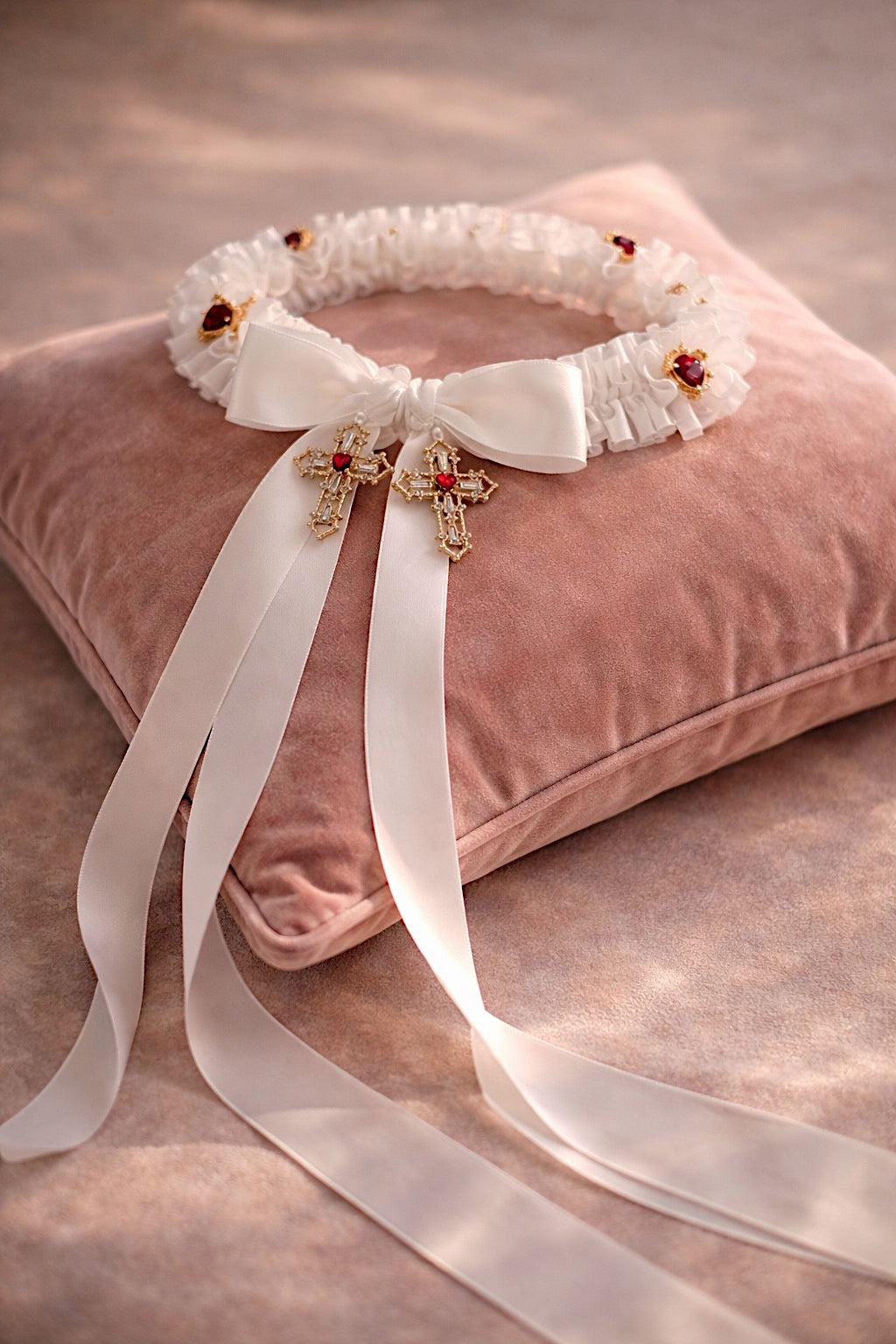 Cross My Heart Jeweled Charm Wedding Garter flat lay with ivory satin ribbons, gold cross charms, and red jeweled heart accents on a blush velvet pillow