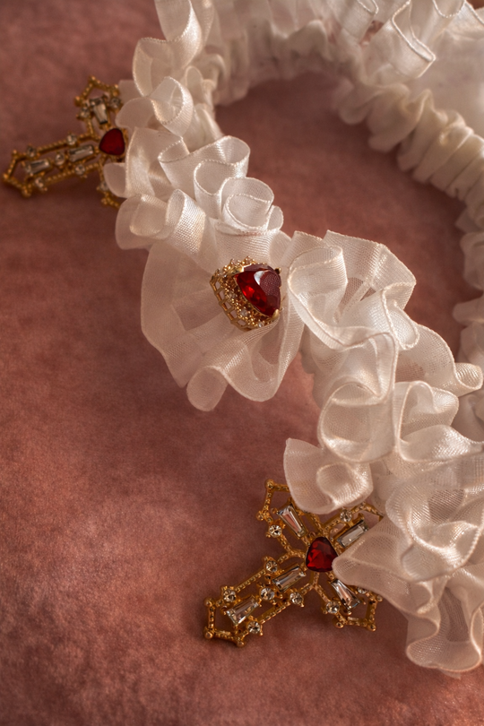 Close-up of the Cross My Heart Jeweled Charm Wedding Garter bow with gold cross charms and red heart jewels on ivory satin ribbon