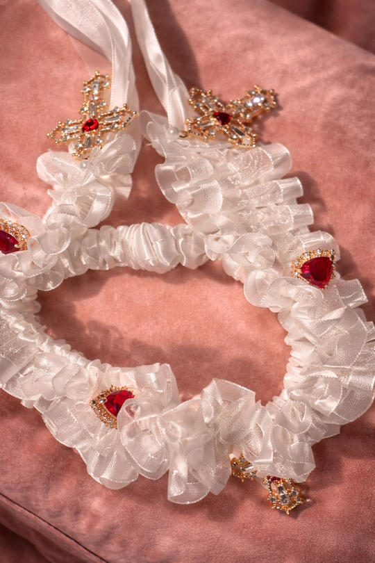 Cross My Heart Jeweled Charm Wedding Garter with ivory ruffles, satin bow, ornate gold cross charms, and red jeweled heart details on a blush velvet pillow