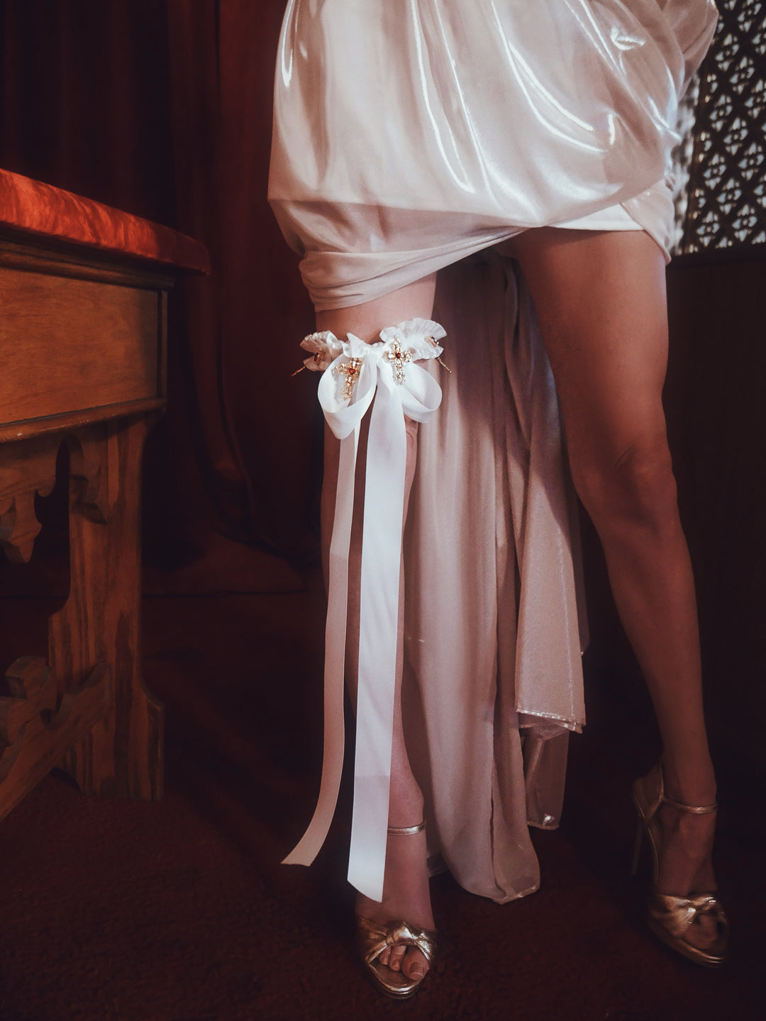 Bride wearing the Cross My Heart Jeweled Charm Wedding Garter with ivory bow and long satin ribbons in a moody chapel-inspired setting