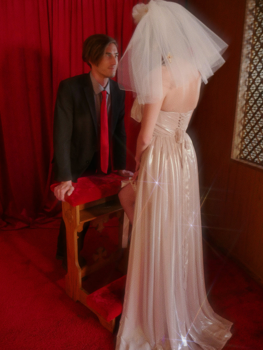 Brunette bride in ivory gown wearing the Cross My Heart Jeweled Charm Wedding Veil while facing a seated man in a chapel-inspired editorial scene