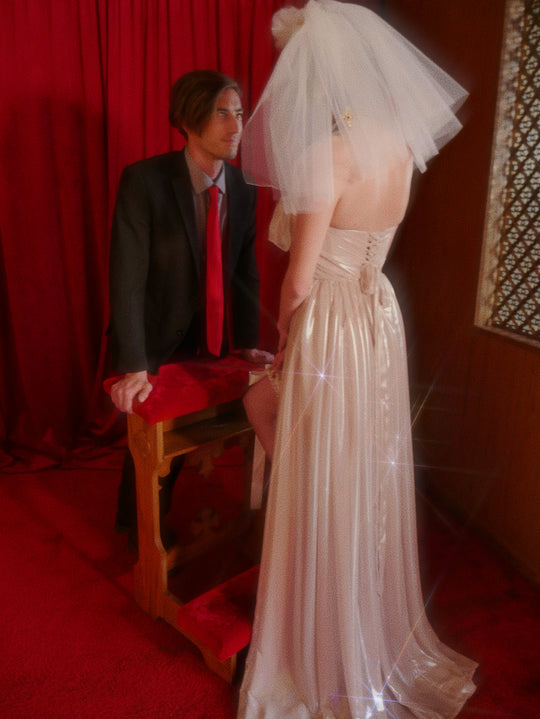 Brunette bride in ivory gown wearing the Cross My Heart Jeweled Charm Wedding Veil while facing a seated man in a chapel-inspired editorial scene