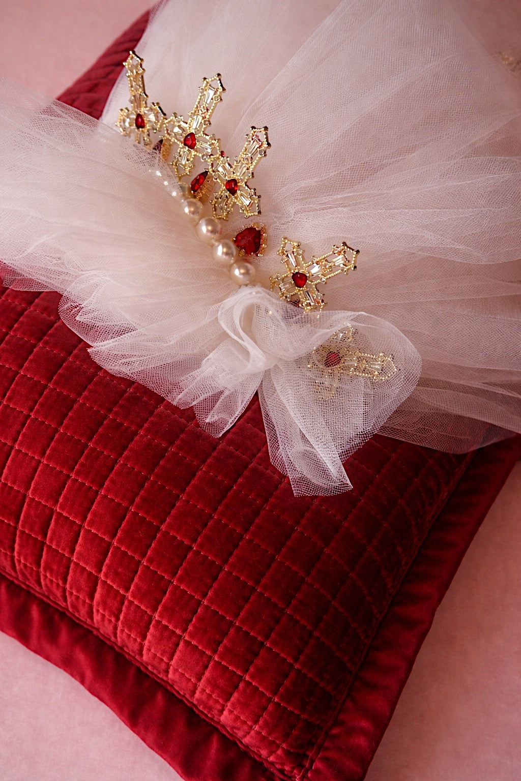 Close-up of the Cross My Heart Jeweled Charm Wedding Veil comb with pearls, ornate gold cross charms, and red jeweled heart details on a red velvet pillow