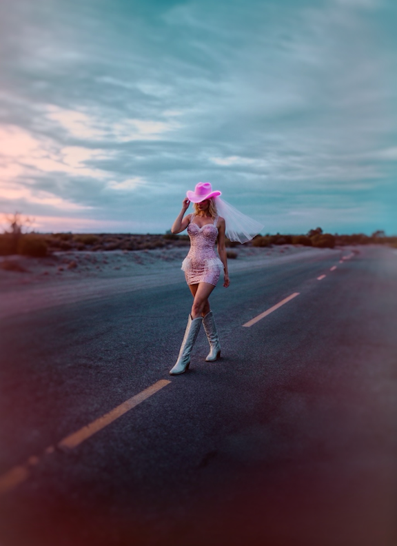 A bride in a glitter mini wedding dress walking on a desert road in cowboy boots and veil.