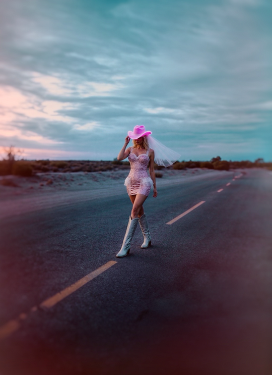 A bride in a glitter mini wedding dress walking on a desert road in cowboy boots and veil.